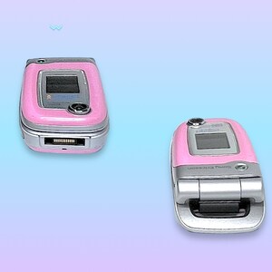 May include: Two pink Sony Ericsson flip phones, one open and one closed, are displayed against a gradient blue and purple background. The phones have silver accents and small screens. The closed phone is on the left, and the open phone is on the right.