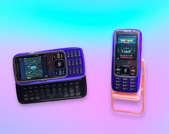 May include: Two purple Samsung mobile phones. One phone is positioned horizontally with a slide-out QWERTY keyboard displaying a Pac-Man game. The second phone is positioned vertically on a small pink folding chair, displaying a Pac-Man Plus game.