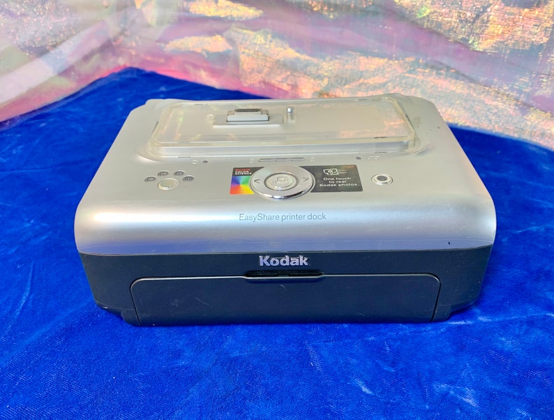 Może przedstawiać: Srebrno-czarna stacja dokująca do drukarki Kodak EasyShare. Drukarka ma przezroczystą plastikową g&oacute;rę i mały kolorowy wyświetlacz. Widoczny jest tekst "EasyShare printer dock" i "Kodak".