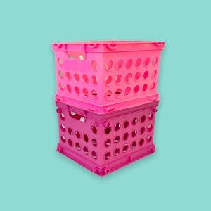 May include: Two stacked, bright pink plastic storage crates. Each crate has a rectangular shape with rounded holes and handles on the sides. The crates are designed for organizing and storage.