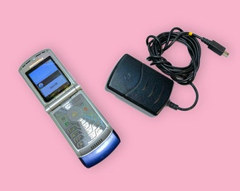 May include: Silver and blue Motorola flip phone with a black charger and cable. The phone is open, displaying a blue screen with the text "Insert SIM". The charger is black with the Motorola logo.