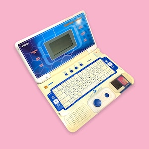 Può includere: Un VTech Genius Notebook vintage, un giocattolo educativo per bambini, è aperto su uno sfondo rosa. Il laptop è principalmente bianco sporco con dettagli blu, un piccolo schermo e una tastiera. La parte superiore del laptop riporta la scritta "Genius Notebook".