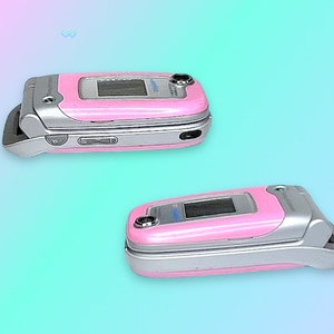 May include: Two pink flip phones with silver accents are displayed against a gradient background. The phones are open and closed, showcasing their screens and external features. The design is reminiscent of early 2000s technology.