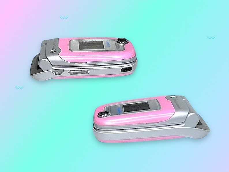 May include: Two pink flip phones with silver accents are displayed against a gradient background. The phones are open and closed, showcasing their screens and external features. The design is reminiscent of early 2000s technology.