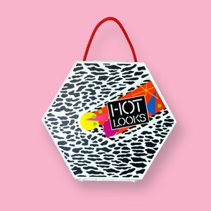 May include: A hexagonal white case with a black and white animal print pattern. The case has a red handle and a sticker that says "HOT LOOKS" with colorful geometric shapes. The background is a solid pink.