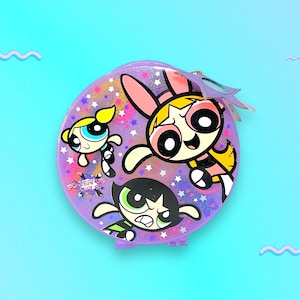 Puede incluir: Un estuche redondo morado con los personajes de dibujos animados de las Supernenas. El estuche tiene un acabado brillante con detalles de estrellas y un dije de mariposa. Las Supernenas están representadas en sus colores característicos: amarillo, rosa y verde.