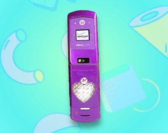 May include: A purple Motorola flip phone featuring a small screen with the Verizon logo, a camera lens, and a large, holographic heart-shaped sticker on the lower section. The device has a metallic finish and displays the Motorola logo above the heart.
