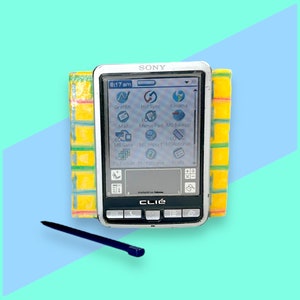 May include: A silver Sony CLIÉ PDA with a stylus, displayed on a light blue and teal background. The screen shows various app icons. The PDA is flanked by a yellow and green patterned object.