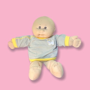 May include: A vintage doll with a bald head and painted facial features. The doll wears a grey long-sleeved top with yellow trim and a white cat graphic. The doll's legs and arms are beige, set against a pink background.