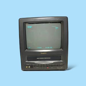 May include: A vintage grey television with an integrated VCR. The screen shows "STOP" and a timer. The VCR has playback buttons and other functions. The brand name "Sansui" is visible on the front.