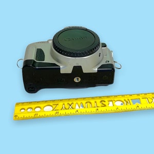 Pode incluir: Uma c&acirc;mera Canon prateada e preta com uma tampa de lente preta. Uma r&eacute;gua amarela com marca&ccedil;&otilde;es pretas e letras recortadas &eacute; colocada ao lado da c&acirc;mera. A c&acirc;mera est&aacute; em um fundo azul claro.