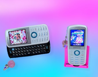 May include: Two silver Samsung cell phones with blue accents are displayed against a gradient background. One phone has a keyboard and a photo on the screen. The other phone is in a pink stand and displays a game. A pink pom-pom charm is attached to the keyboard phone.
