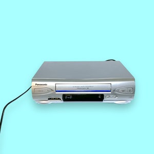 May include: Silver Panasonic VCR player on a light blue background. The VCR has a black power cord and features buttons and a slot for video tapes. The front of the VCR has the Panasonic logo.
