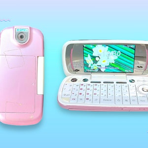 Peut inclure: Un téléphone à clapet rose et blanc avec un clavier complet et un écran affichant un fond d'écran floral et l'heure 12h30. Le téléphone est ouvert, révélant l'écran et le clavier, et fermé, montrant l'appareil photo et le compartiment de la batterie.