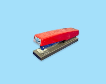 May include: A red, blue, and black stapler with the text "ETONA-21" on the side. The stapler is on a light blue background. The stapler is a common office supply.