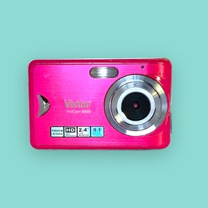 May include: A bright pink Vivitar ViviCam 8025 digital camera. The camera has a touch screen, HD, a 2.4" LCD monitor, and 8.1 megapixels. The camera has a silver lens and flash. The camera is on a light blue background.