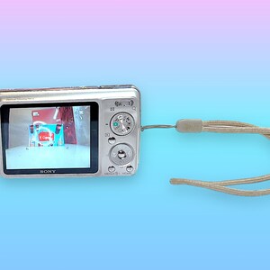 May include: A silver Sony digital camera with a screen displaying an image. The camera has several buttons and a beige wrist strap. The background is a gradient of blue and pink.