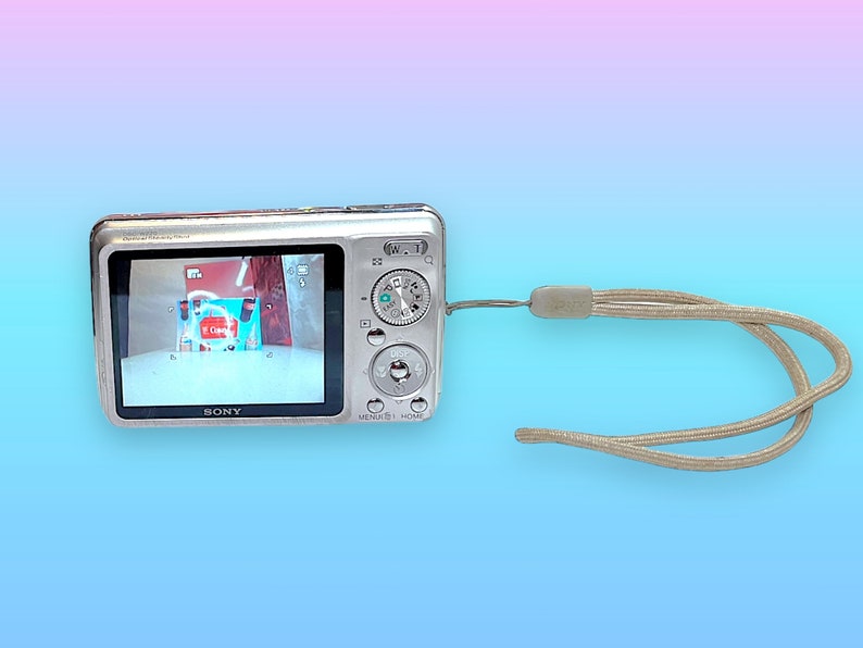May include: A silver Sony digital camera with a screen displaying an image. The camera has several buttons and a beige wrist strap. The background is a gradient of blue and pink.