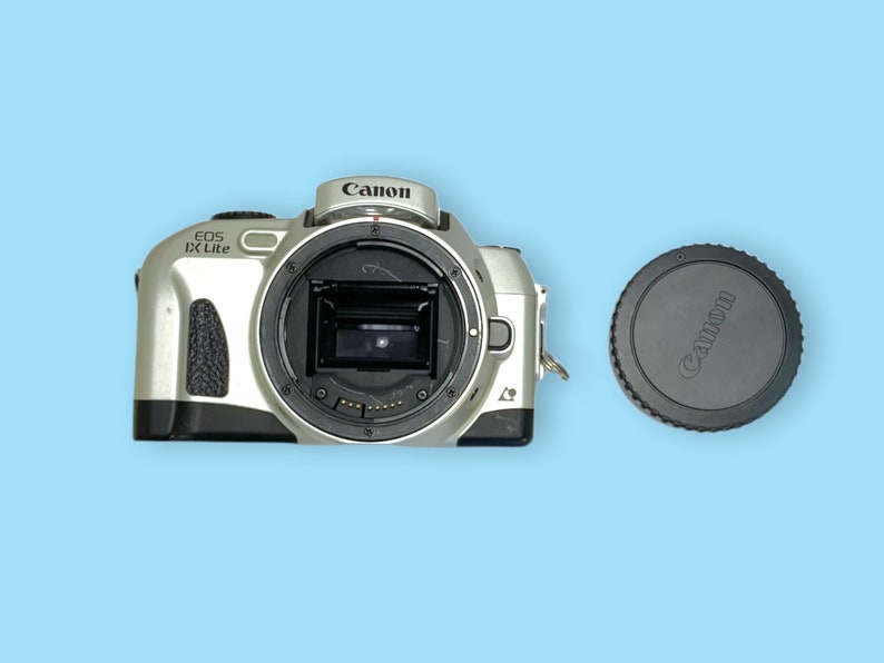 Pode incluir: Corpo de c&acirc;mera Canon EOS IX Lite prateado com uma tampa de lente preta. O corpo da c&acirc;mera tem o logotipo da Canon e "EOS IX Lite" impresso. A tampa da lente tamb&eacute;m tem o logotipo da Canon. Em um fundo azul claro.