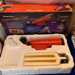 May include: A vintage Fisher-Price Starscope Set for kids. The red telescope is mounted on a white tripod. The box is orange and blue with the product name and images of space. The set includes pre-programmed space images.