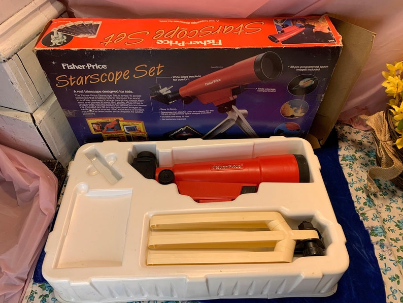 May include: A vintage Fisher-Price Starscope Set for kids. The red telescope is mounted on a white tripod. The box is orange and blue with the product name and images of space. The set includes pre-programmed space images.
