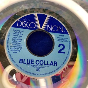 May include: A vintage MCA DiscoVision videodisc for "Blue Collar." The disc features a blue and white label with the film title and the number 2. The disc is encased in a clear plastic case, with the text "Warning: All rights reserved" visible.