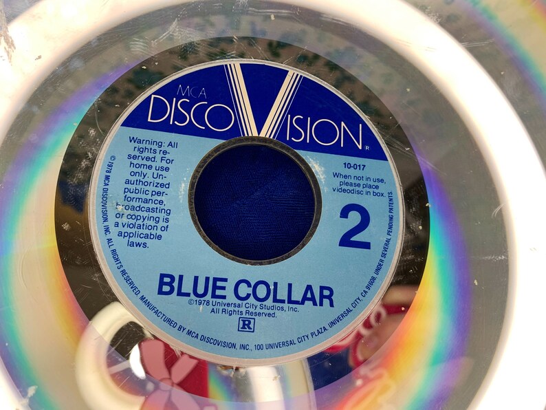 May include: A vintage MCA DiscoVision videodisc for "Blue Collar." The disc features a blue and white label with the film title and the number 2. The disc is encased in a clear plastic case, with the text "Warning: All rights reserved" visible.