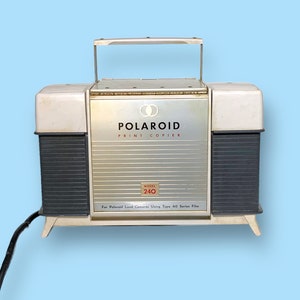 May include: Vintage Polaroid Print Copier Model 240. The device has a silver and white body with black accents and a handle. The Polaroid logo and model number are visible on the front. It is designed for Polaroid Land Cameras using Type 40 Series film.