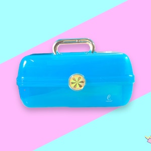 Puede incluir: Un estuche de maquillaje azul brillante de estilo vintage con un asa plateada y un cierre circular dorado. El estuche tiene un acabado brillante y la letra "c" está impresa en la esquina inferior derecha. El fondo es una división de rosa y azul claro.