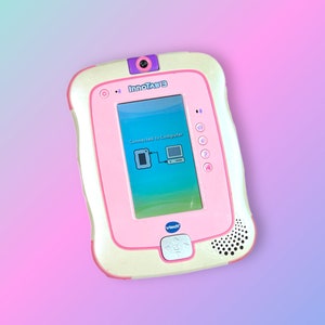 Può includere: Un tablet educativo VTech InnoTab 3 rosa e crema. Lo schermo mostra un grafico di connessione. Il tablet ha una fotocamera, un altoparlante e diversi pulsanti. Il testo "Connected to Computer" è visibile.