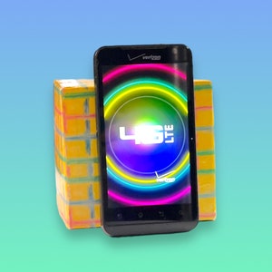 Op de afbeelding: Een zwarte smartphone met het Verizon-logo en een kleurrijke 4G LTE-afbeelding op het scherm. De telefoon staat tegen een gele en veelkleurige blok.