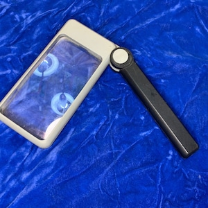 May include: A rectangular, handheld magnifying glass with a white frame and a black handle. The lens is clear, and the handle is folded at an angle. The background is a textured, deep blue fabric.