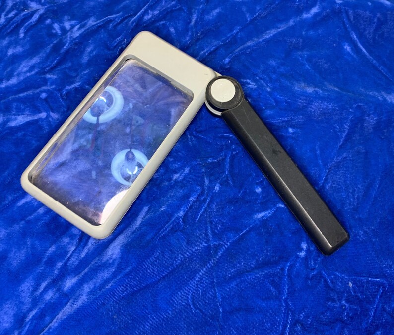 May include: A rectangular, handheld magnifying glass with a white frame and a black handle. The lens is clear, and the handle is folded at an angle. The background is a textured, deep blue fabric.