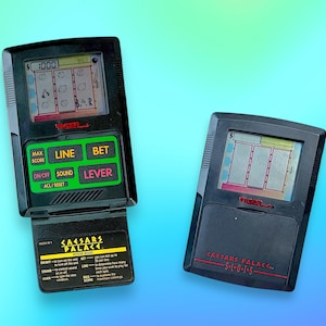 Op de afbeelding: Twee zwarte draagbare elektronische gokautomaten. Eén is open en toont een scherm met gokautomaatgraphics en de tekst "$1000". De andere is gesloten. Beide hebben de tekst "CAESARS PALACE" en "TIGER" erop.