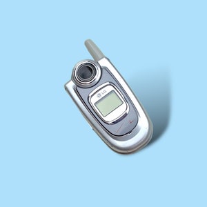 Könnte beinhalten: Silbernes LG-Klapphandy mit kleinem Bildschirm und Antenne, mit Lautsprecher und Verizon-Logo. Das Telefon befindet sich auf einem hellblauen Hintergrund.