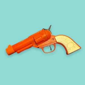 Op de afbeelding: Fel oranje speelgoedpistool met een crèmekleurig handvat, tegen een lichtblauwe achtergrond. Het speelgoedpistool heeft een klassiek revolverontwerp met ingewikkelde details op de loop en het lichaam. De handgreep heeft een decoratieve ovale vorm.