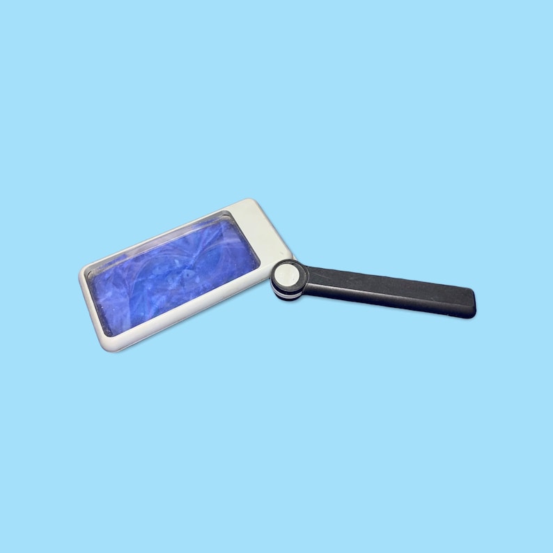 May include: A rectangular magnifying glass with a white frame and a black handle. The lens has a blue, marbled appearance. The handle is connected to the frame with a circular joint. The background is a solid light blue.