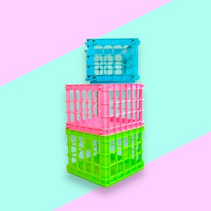 Puede incluir: Tres cajas de almacenamiento de plástico de colores apiladas una encima de la otra. Las cajas son de color verde brillante, rosa y azul, con un diseño de rejilla. El fondo es una división diagonal de rosa y azul claro.