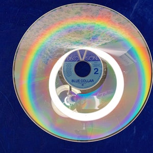 May include: A vintage Discovision disc with a rainbow-like reflection. The disc features the text "Blue Collar" and the number "2". The image is set against a dark blue background, creating a striking contrast.