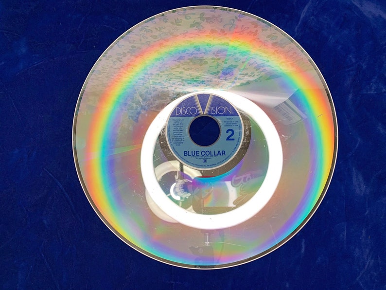 May include: A vintage Discovision disc with a rainbow-like reflection. The disc features the text "Blue Collar" and the number "2". The image is set against a dark blue background, creating a striking contrast.