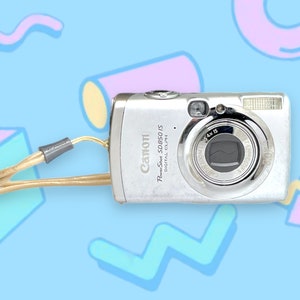 May include: Silver Canon PowerShot SD850 IS digital camera with a tan wrist strap. The camera has a lens, flash, and various buttons. The background is light blue with colorful geometric shapes.