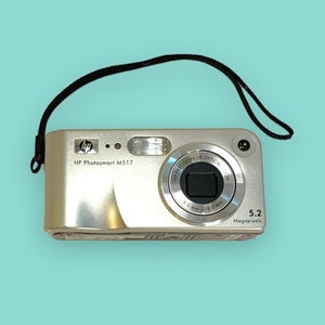 Op de afbeelding: Een zilveren HP Photosmart M517 digitale camera met een zwart polsbandje. De camera heeft een lens, flitser en de tekst "5.2 Megapixels". De camera staat op een lichtblauwe achtergrond.