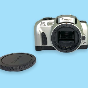 Pode incluir: C&acirc;mera Canon EOS IX Lite prateada com montagem de lente preta e tampa de lente preta. A c&acirc;mera tem o logotipo da Canon na parte superior e est&aacute; em um fundo azul claro.