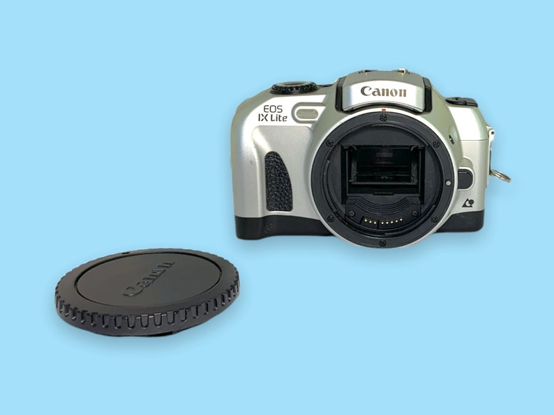 Pode incluir: C&acirc;mera Canon EOS IX Lite prateada com montagem de lente preta e tampa de lente preta. A c&acirc;mera tem o logotipo da Canon na parte superior e est&aacute; em um fundo azul claro.
