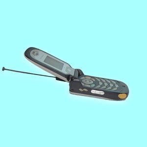 Peut inclure: Un téléphone à clapet noir et argenté avec une antenne déployée. Le téléphone est ouvert, révélant un écran et un clavier. Le téléphone a un accent doré sur le côté, sur fond bleu clair.