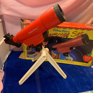 May include: A red Fisher-Price Starscope Set toy telescope with a white tripod. The telescope is angled towards the left, with the box in the background. The box has an image of the telescope and the words "Starscope Set".