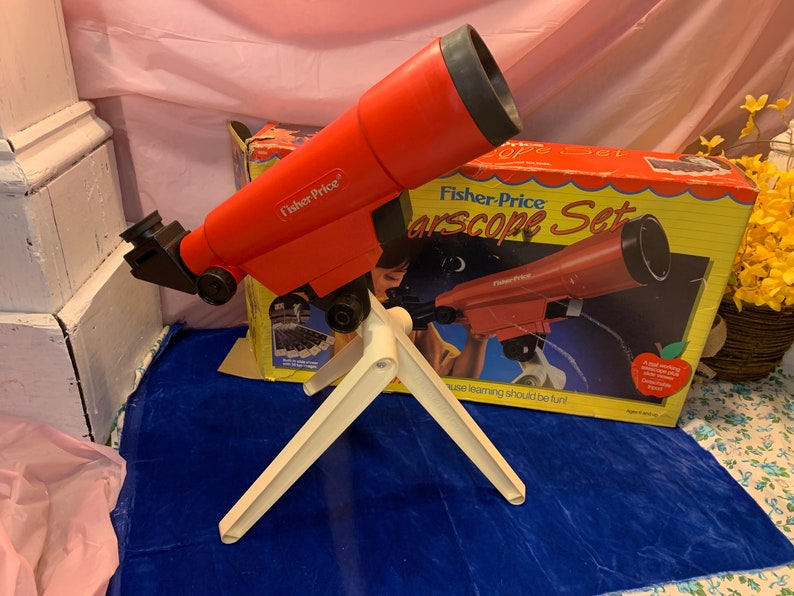 May include: A red Fisher-Price Starscope Set toy telescope with a white tripod. The telescope is angled towards the left, with the box in the background. The box has an image of the telescope and the words "Starscope Set".