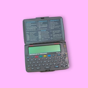 Può includere: Un dispositivo elettronico vintage nero Dizionario & Thesaurus con uno schermo verde e una tastiera QWERTY completa. Il dispositivo ha un coperchio ribaltabile con istruzioni. Il testo "DICTIONARY & THESAURUS" è stampato sul dispositivo.