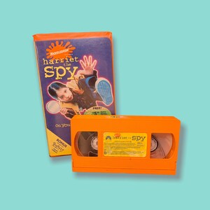 May include: A Nickelodeon Harriet the Spy VHS tape and its matching orange plastic case. The cover features a purple background with a girl holding a magnifying glass, the movie title, and a bonus Rugrats music video label. The tape is bright orange.