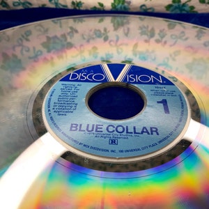May include: A vintage DiscoVision disc with the title "Blue Collar." The disc has a reflective surface with rainbow hues. The label is blue and white, with the DiscoVision logo and copyright information.